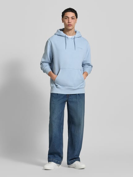 Męski model nosi Hoodie mit Label-Stitching und Känguru-Tasche von Dickies, bawełniany miks, krój relaxed fit, jasnoniebieski kolor, cała sylwetka z przodu.