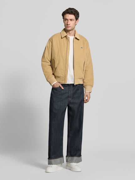 Männermodel trägt Bomberjacke mit Cordstruktur von LACOSTE in Beige mit Strukturmuster, Reißverschluss und Logo-Patch, Ganzkörperansicht von vorne.