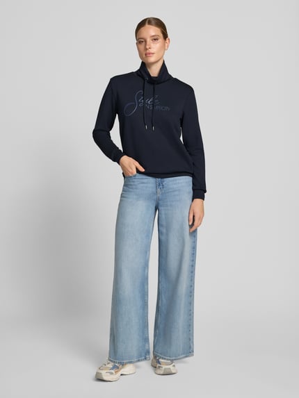 Frauenmodel trägt Regular Fit Sweatshirt aus reiner Modal-Mix Modell 'BANU' von Soyaconcept in Dunkelblau mit hellblauem Schriftzug, kombiniert mit weiter Jeans und Sneakers, Damen Sweatshirt von Soyaconcept, Ganzkörperansicht von vorne.