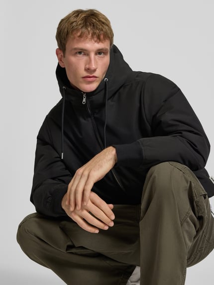 Męski model nosi Jacke mit Kapuze und Eingrifftaschen Modell 'TRACE' od JACK & JONES, czarna, krój regular fit, z kapturem i kieszeniami, zdjęcie z przodu, zbliżenie na górną część ciała.