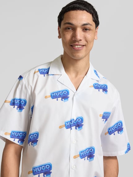 Mężczyzna nosi Relaxed Fit Freizeithemd aus reiner Baumwolle Modell 'ELIGINO' od Hugo Blue, białe z niebieskim wzorem, krój relaxed fit, resortkragen, krótki rękaw, guziki, widok na górną część ciała z przodu.
