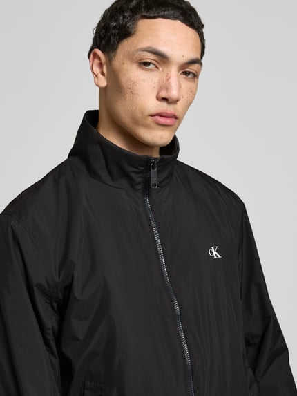 Männermodel trägt schwarze Trainingsjacke mit Logo und Stehkragen von Calvin Klein Jeans, Nahaufnahme Oberkörper, Regular Fit, Reißverschluss.