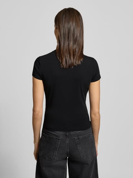 Kobieta modelka nosi Slim Fit T-Shirt mit Allover-Strasssteinbesatz od Guess, damski t-shirt, stójka, krój slim fit, obszycie z kamieni stras na całej powierzchni, detal z logo, mieszanka bawełny i elastanu, długość tyłu 51 cm dla rozmiaru S, czarny kolor, gładka faktura, widok całej sylwetki z tyłu.