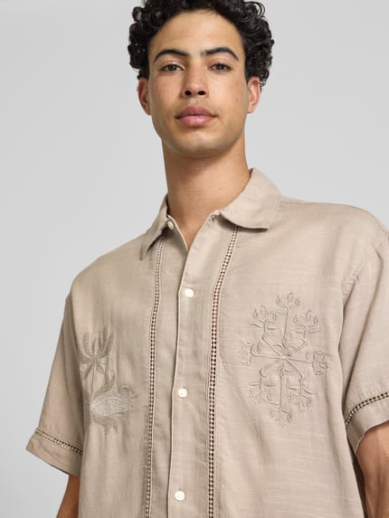 Męski model nosi Relaxed Fit Freizeithemd aus Lyocell-Mix Modell 'Menton' od Jack & Jones z ażurowymi haftami, mieszanka bawełny i lyocellu, krój relaxed fit, z krótkim rękawem, kołnierzyk typu kent, listwa guzikowa, florystyczny haft, długość tyłu w rozmiarze M 72 cm, szerokość w rozmiarze M 52 cm, długość rękawów w rozmiarze M 25 cm, jasnobeżowy materiał, lekko teksturowana tkanina, widok z przodu, zbliżenie na górną część ciała