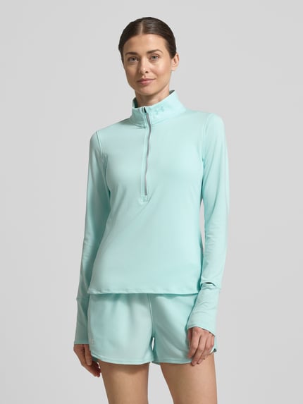 Frauenmodel trägt Longsleeve mit Stehkragen und Reißverschluss von Under Armour in Hellblau, glatte Oberfläche, Regular Fit, Ganzkörperansicht von vorne.