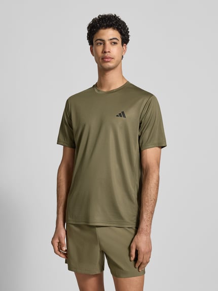 Männermodel trägt T-Shirt mit Label-Print von adidas Training in Olivgrün, Lockerer-Schnitt, gerippter Rundhalsausschnitt, Nahaufnahme Oberkörper. Rückenlänge bei Größe M: 70 cm.