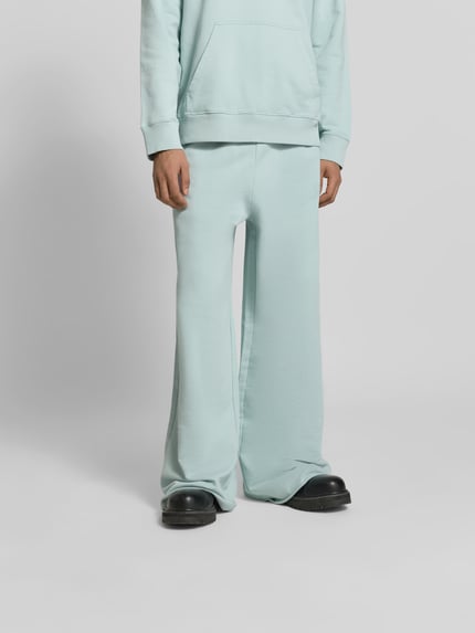 Męski model nosi spodnie dresowe Wide Fit Sweatpants mit elastischem Bund von MM6 Maison Margiela, czysta bawełna, krój wide fit, wyhaftowane logo, kieszeń z tyłu, długość wewnętrzna nogawki dla rozmiaru M: 84 cm, szerokość w rozmiarze M: 41 cm, kolor jasnoniebieski, tekstura miękka, gładka powierzchnia, całkowity widok z przodu.