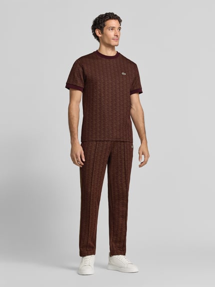 Männermodel trägt Regular Fit T-Shirt aus Baumwoll-Mix von Lacoste in Dunkelbraun mit Muster und weißem Logo, Ganzkörperansicht von vorne.