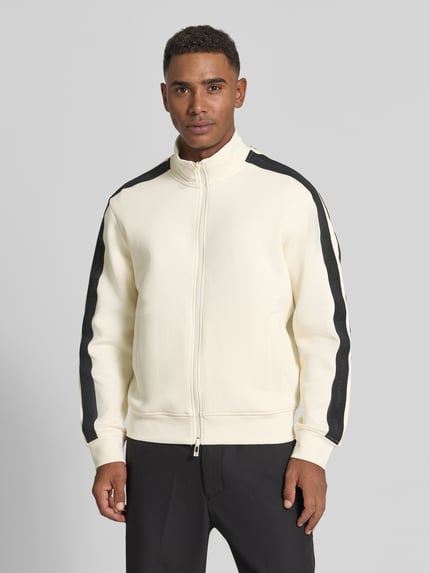 Męski model nosi Regular Fit Sweatjacke aus Baumwoll-Mix Modell 'Felpa' od Armani Exchange w kolorze kremowym z czarnymi lampasami na rękawach, wpuszczane kieszenie, stójka, zamek błyskawiczny dwustronny, elastyczne wykończenia, Długość tyłu w rozmiarze M: 64 cm, Długość rękawów w rozmiarze M: 62 cm, Ganzkörperansicht von vorne.