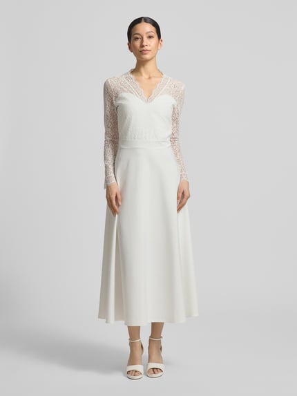 Frauenmodel trägt Tailliertes Brautkleid mit V-Ausschnitt Modell 'Joanna' von GUIDO MARIA KRETSCHMER WOMAN aus Elastischer-Material-Mix, Langärmelig, Oberteil aus Spitze, blickdichter Rockteil, Nahtreißverschluss, Hochzeitsmode, Ganzkörperansicht von vorne.