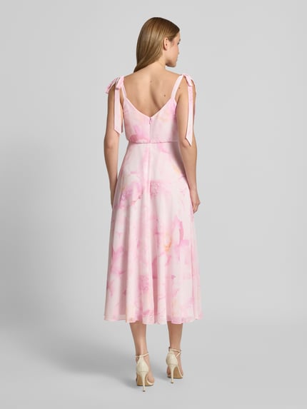 Vrouwenmodel draagt knielange cocktailjurk met bloemen all-over print van Jake*s Cocktail, getailleerde pasvorm, onderrok, mouwloos design, sierstrikken, ruglengte maat 36: 102 cm, borstwijdte maat 36: 40 cm, lichtroze tinten, vloeiende stof, geheelbeeld achteraan.