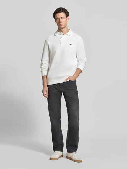 Männermodel trägt weißes Regular Fit Sweatshirt aus reiner Baumwolle von Lacoste zu dunkler Jeans, Ganzkörperansicht von vorne. Herren Sweatshirt von Lacoste