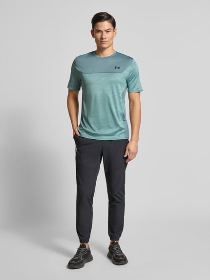 Männermodel trägt Funktionsshirt mit Logo und Rundhalsausschnitt von Under Armour, Material-Mix mit Stretch-Anteil, Regular-Fit, grün-blau, schwarze Hose, Rückenlänge bei Größe M 71 cm, Ganzkörperansicht von vorne.