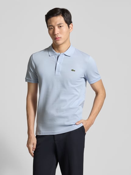 Männermodel trägt ein Slim Fit Poloshirt aus reiner Baumwolle von Lacoste in Hellblau mit Logo-Stitching, Piqué-Struktur, Ganzkörperansicht von vorne.