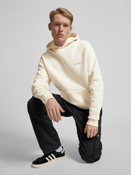 Männermodel trägt Hoodie mit Label-Print von REVIEW in Creme, Baumwoll-Mix, gerade Form, Känguru-Tasche, Nahaufnahme Oberkörper.