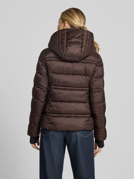 Frauenmodel trägt Steppjacke mit Kapuze und Reißverschlusstaschen von MICHAEL Michael Kors in Dunkelbraun, gesteppte Textur, Ganzkörperansicht Rückansicht. Modell 'PACKABLE WITH BIB', gerader Schnitt, Kapuze, seitliche Reißverschlusstaschen, elastische Abschlüsse mit Daumenloch.