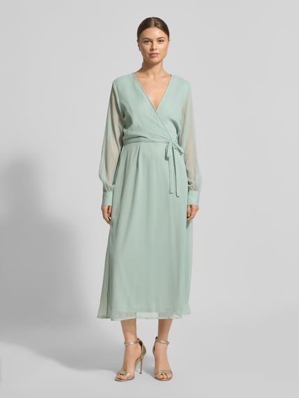 Frauenmodel trägt Midikleid mit Taillenband Modell 'FALIA' von Vila in Hellgrün, Wickel-Optik, V-Ausschnitt, semitransparente Optik, langärmlig, blickdichter Unterrock, taillierte Passform, Ganzkörperansicht von vorne.
