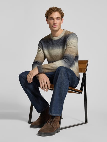 Männermodel trägt Strickpullover mit Rundhalsausschnitt Modell 'AIDEN' von Samsøe Samsøe, Farbverlauf in Beige und Blau, Material-Mix mit Alpaka-Anteil, gerade geschnitten, kombiniert mit Jeans und braunen Schnürstiefeln, Ganzkörperansicht von vorne.