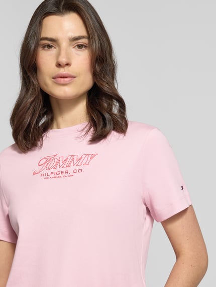 Vrouwenmodel draagt Slim Fit T-Shirt aus reiner Baumwolle van Tommy Hilfiger in lichtroze, uni, label print, ronde hals, korte mouwen, jersey, puur katoen. Close-up bovenlichaam.