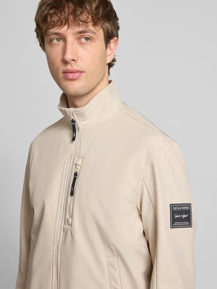 Männermodel trägt Regular Fit Bomberjacke mit Stehkragen Modell 'Parker' von Jack & Jones in Beige, Softshell, Nahaufnahme Oberkörper. Kontext: Herrenjacke mit Napoleontasche, Eingrifftaschen mit Druckknopfverschluss, elastische Abschlüsse, Maße bei Größe M: Rückenlänge 67 cm, Schulterbreite 43 cm, Brustweite 55 cm, Ärmellänge 65 cm.