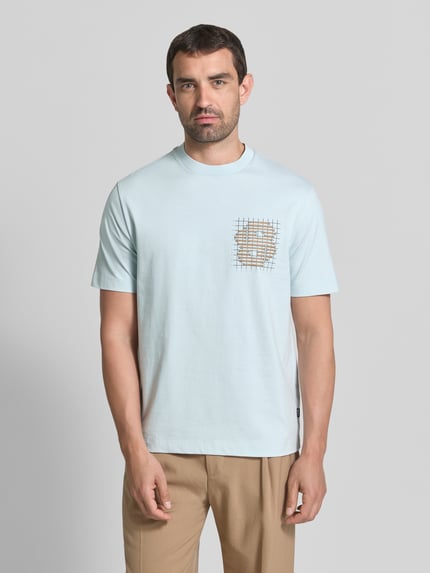 Mężczyzna nosi jasnoniebieski t-shirt model 'C-Tigerman 49' z bawełny od BOSS, beżowe spodnie, z przodu nadruk; Ganzkörperansicht von vorne. Męski t-shirt od BOSS.