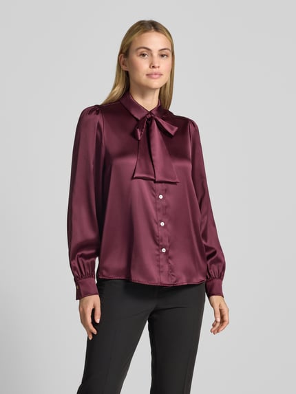 Vrouwenmodel draagt Regular Fit Hemdbluse aus Satin van Christian Berg Woman Selection, bordeauxrood met glanzende satijntextuur, sierstrik, hemdkraag, lange mouwen, voorkant volledig zichtbaar, Ganzkörperansicht von vorne.