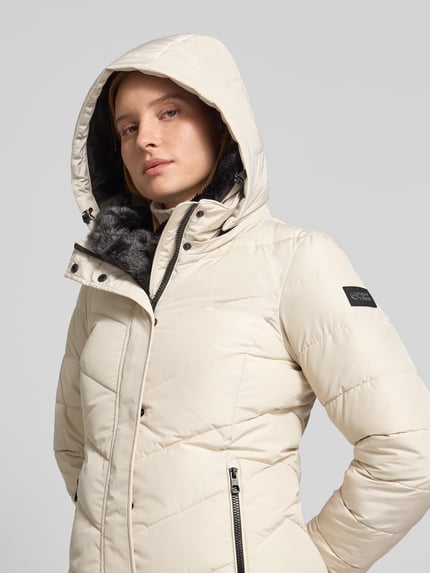 Frauenmodel trägt Steppmantel mit Kapuze von NAUTGÅRD 'NEBULA' in Creme mit Reißverschlusstaschen, Nahaufnahme Oberkörper, Regular Fit.