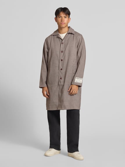 Männermodel trägt Trenchcoat mit Knopfleiste und Label-Patch von ANOTHER COTTON LAB, braun kariert, gerade geschnitten, Ganzkörperansicht von vorne.
