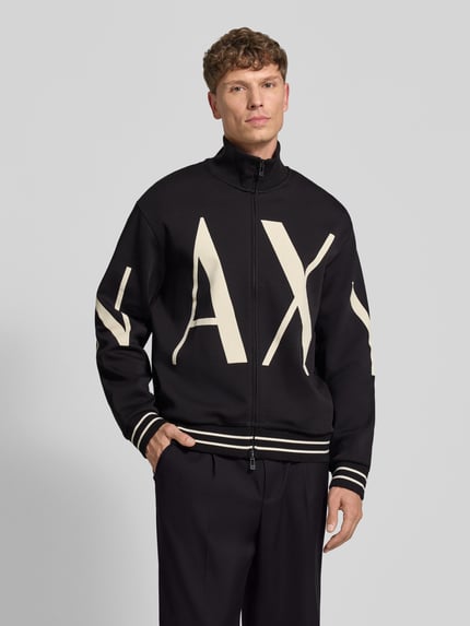 Męski model nosi Sweatjacke aus Baumwoll-Mix von Armani Exchange, czarne tło z białym nadrukiem, luźny fason, dzianinowe ściągacze, dwukierunkowy zamek, nadruk label, długość tyłu w rozmiarze M 67 cm, długość rękawa 63 cm, całkowity widok z przodu.