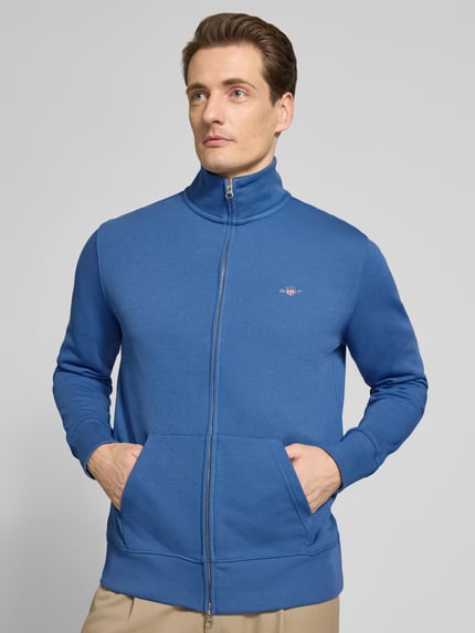 Männermodel trägt Regular Fit Sweatjacke mit Stehkragen Modell 'Gield' von Gant, blau, mit Logo-Stitching, Ganzkörperansicht von vorne. Baumwoll-Mix, Reißverschluss, geteilte Kängurutasche, elastische Abschlüsse.