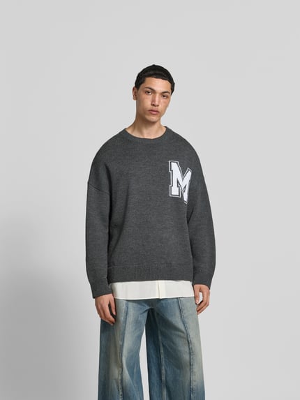Męski model nosi Oversized Pullover aus Woll-Mix od MM6 Maison Margiela, kolor szary, imitacja dzianiny, wyhaftowane logo, okrągły dekolt, długość tyłu dla rozmiaru M: 80 cm, krój oversized fit, widok całej sylwetki z przodu.