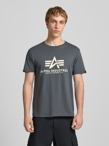 Männermodel trägt T-Shirt mit Logo und Rundhalsausschnitt von Alpha Industries, reine Baumwolle, Regular Fit, grauer Farbton, Ganzkörperansicht von vorne