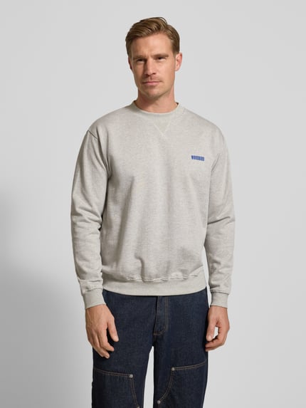 Männermodel trägt Sweatshirt mit Label-Print Modell 'Cane' von WOODBIRD in Hellgrau, Rundhalsausschnitt, gerippte Abschlüsse, Ganzkörperansicht von vorne.