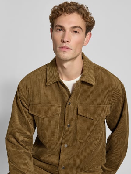 Męski model nosi Regular Fit Hemdjacke aus Cord Modell 'David' von Wood Wood, brązowy sztruks, kołnierzyk typu kent, kieszenie na piersi, bawełna, krój regular fit, długość tyłu w rozmiarze M: 73 cm, zdjęcie w zbliżeniu na górną część ciała.