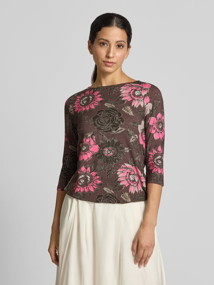 Modelka nosi T-Shirt im Allover-Look mit 3/4-Arm von Christian Berg Woman, mieszanka wiskozy i elastanu, krój slim fit, wzór na całej powierzchni, okrągły dekolt, rękaw o dł. 3/4, Długość tyłu w rozmiarze S: 55 cm, brązowe tło, różowe i beżowe kwiaty, prosta faktura, Ganzkörperansicht von vorne.