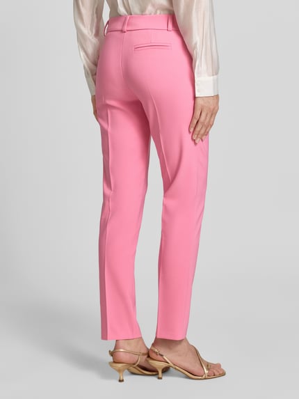 Modelka nosi Stoffhose mit Bügelfalte Modell 'REWI' od Rinascimento, różowy materiał z prasowanym kantem, krój slim fit, widok całej sylwetki z tyłu. Szerokość w rozmiarze S: 40 cm, długość nogawki od wewnątrz: 75 cm, domieszka stretchu, francuskie kieszenie, imitacja tylnej kieszeni.