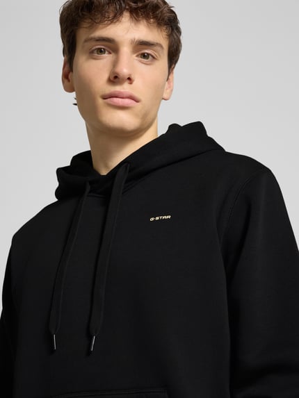 Männermodel trägt Hoodie mit Kapuze von G-Star Raw in Schwarz, locker geschnitten, Label-Print, Nahaufnahme Oberkörper.