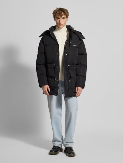 Męski model nosi Parka mit Kapuze Modell 'Briska' von Don´t Waste Culture, czarny, luźny krój, bawełniana tekstura, przód z zakrytym zamkiem, nadruk z logo, kieszenie z patkami, długość tyłu w rozmiarze M 81 cm, szerokość ramion w rozmiarze M 60 cm, szerokość klatki piersiowej w rozmiarze M 63 cm, długość rękawa w rozmiarze M 60 cm, pełny widok z przodu.
