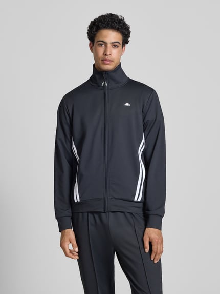 Männermodel trägt Sweatjacke mit Reißverschlusstaschen Modell 'TOSCIANA' von Ellesse, Schwarz, Material-Mix mit Stretch-Anteil, Ganzkörperansicht von vorne.