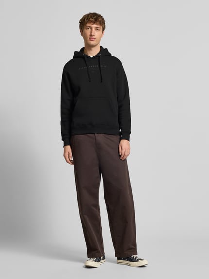 Männermodel trägt Relaxed Fit Hoodie aus Baumwoll-Mix mit Label-Detail von Alpha Industries in Schwarz, Känguru-Tasche, Kapuze, Ganzkörperansicht von vorne.