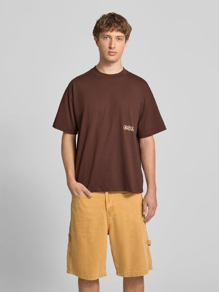 Mężczyznę model nosi t-shirt mit Label-Stitching und Rundhalsausschnitt von ANOTHER COTTON LAB, bawełna, krój relaxed fit, długość tyłu w rozmiarze M 69 cm, brązowy kolor, gładka struktura. Ganzkörperansicht von vorne.
