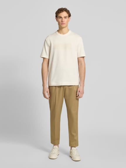 Męski model nosi T-Shirt z logo i okrągłym dekoltem od ARMANI EXCHANGE, czysta bawełna, krój regular fit, kolor kremowy, widok całej sylwetki z przodu. Długość pleców w rozmiarze M: 71 cm.