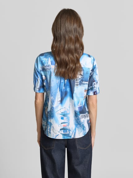 Kobiece model prezentuje Regular Fit Hemdbluse mit Allover-Motiv-Print von Marc Cain z bawełny, niebieski nadruk, kołnierzyk koszulowy, krótkie rękawy, elastyczne zakończenia rękawów, długość tyłu w rozmiarze 36: 62 cm, długość rękawów w rozmiarze 36: 28 cm, widok całej sylwetki z tyłu.