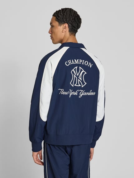Männermodel trägt Relaxed Fit Tracksuit mit Logo-Patch von Champion in Dunkelblau und Weiß, Stehkragen, Rückenansicht, Kontext Herren Tracksuit, elastische Abschlüsse, Reißverschluss, Bundweite 39 cm, Rückenlänge 68 cm, Schulterbreite 42 cm, Ärmellänge 49 cm, Innenbeinlänge 72 cm. Ganzkörperansicht Rückansicht.