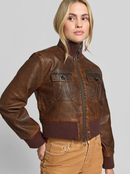 Frauenmodel trägt Lederjacke Modell 'BAILEY' mit Stehkragen von BAUM & PFERDGARTEN in brauner Lederoptik mit Struktur, Ziernähte, metallische Akzente, Zierknöpfe, Label-Detail, Regular Fit, cropped, gerippte Abschlüsse, Zweiwege-Reißverschluss, Brusttasche, Eingrifftaschen, Nahaufnahme Oberkörper.