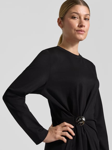 Frauenmodel trägt Midikleid mit Drapierung Modell 'CAMESSIA' von DRYKORN in Schwarz, glatte Textur, Nahaufnahme Oberkörper. Taillierter Schnitt, Rundhalsausschnitt, lange Ärmel, dekorative Schnalle, Kontext: Rückenlänge 121 cm, Brustweite 35 cm, Ärmellänge 59 cm.