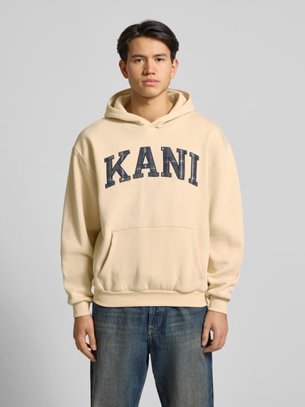 Männermodel trägt Hoodie mit Label-Stitching und Känguru-Tasche von KARL KANI, Oversized, beige, Baumwoll-Mix, Ganzkörperansicht von vorne.