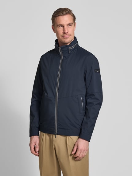 Männermodel trägt Regular Fit Jacke mit Stehkragen von bugatti in Dunkelblau, mit Zweiwege-Reißverschluss, Logo-Detail, Eingrifftaschen, elastischen Ärmelabschlüssen. Rückenlänge bei Größe 50: 68 cm, Schulterbreite: 42 cm, Brustweite: 50 cm, Ärmellänge: 65 cm. Ganzkörperansicht von vorne.