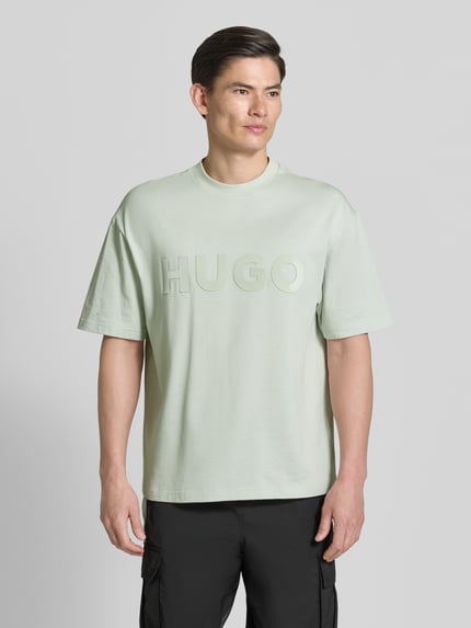 Männermodel trägt Relaxed Fit T-Shirt aus reiner Baumwolle Modell 'DINKEE' von HUGO in Optik: Unifarben mit 3D Label-Print, Rundhals-Ausschnitt, Ärmelform: Kurzarm, Materialverarbeitung: Jersey, Nahaufnahme Oberkörper.