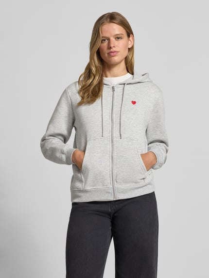 Frauenmodel trägt graue Relaxed Fit Sweatjacke aus Baumwoll-Mix Modell 'CHILLI' von PIECES, rote Stickerei, Ganzkörperansicht von vorne.